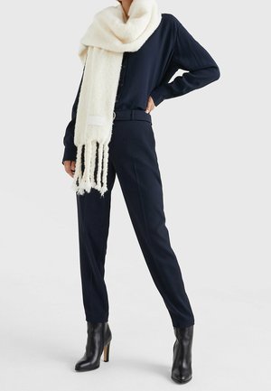 Personne portant une chemise noire boutonnée rentrée dans un pantalon noir assorti, des bottes noires à talons et une longue écharpe blanche à franges.