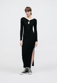 Rochie neagră cu mâneci lungi, cu dungi și decupaj în zona gâtului, cu tăieturi pe lateral, combinată cu adidași negri înalți. Textură netedă și croială mulată.