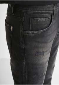 Jeans de mezclilla negra con un acabado desgastado, que presentan áreas rasgadas, una etiqueta en el bolsillo trasero y un diseño ligeramente tapered. Detalles de costura acentuados.