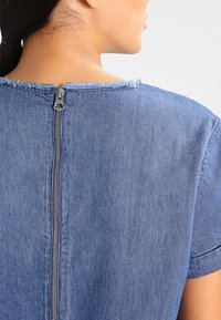Denimová blůza s nezačištěným výstřihem, krátkými rukávy a viditelným zipem na zádech. Tkanina je měkká a modrá s jemnou texturou.