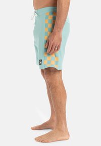 Tyrkysové plavky s bočným vzorom žltého šachovnicového motívu. Materiál sa zdá byť ľahký s viazaním v páse a zaobleným lemom.