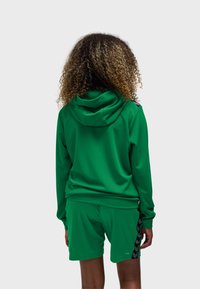 Person med krøllet hår iført en grøn hættetrøje og matchende grønne shorts med sorte chevronstriber, set bagfra mod en hvid baggrund.