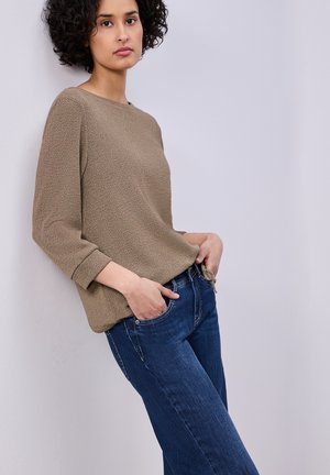 Femme aux cheveux bouclés portant un pull marron et un jean bleu, appuyée contre un mur gris clair, les mains dans les poches.