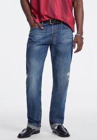 Jeans desgastados azules con un corte relajado, ligero desvanecimiento y acentos deshilachados, combinados con una camisa de rayas rojas y un cinturón negro.