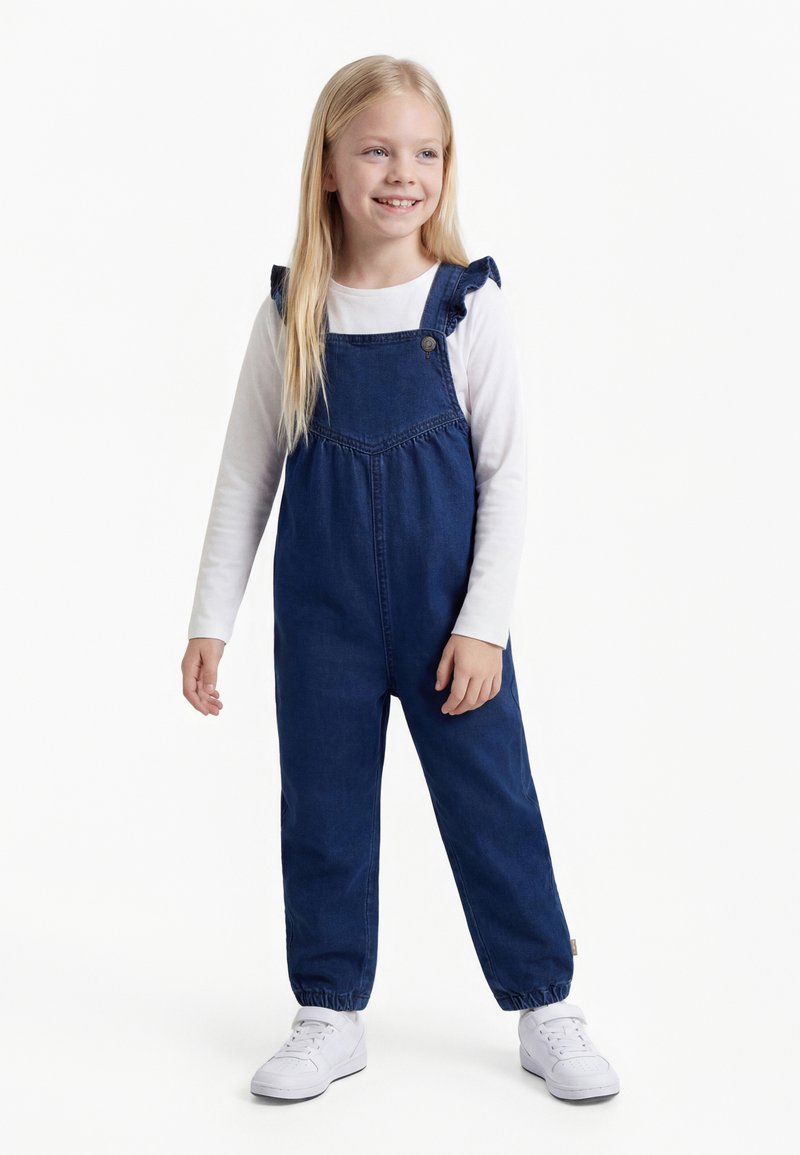 Fille souriante aux longs cheveux blonds portant une chemise blanche à manches longues, une salopette en jean bleu avec des bretelles à volants, et des baskets blanches, debout.