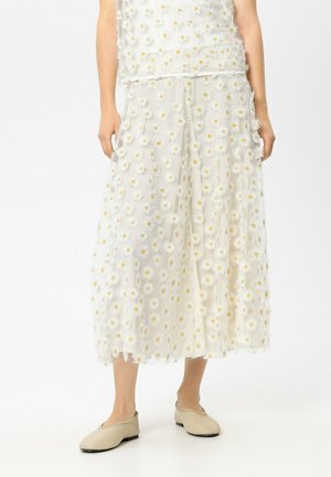 Weißer Midirock mit transparentem Überwurf, verziert mit kleinen weißen und gelben Gänseblümchen, kombiniert mit beigen flachen Schuhen vor hellem Hintergrund.