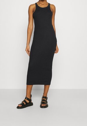 Femme portant une robe midi noire ajustée et sans manches, avec des sandales plates noires à lanières, debout contre un mur uni.