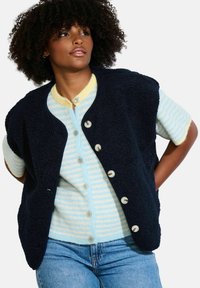 Gilet in pile blu navy con scollatura arrotondata e cinque bottoni, indossato sopra una maglietta a maniche corte a righe blu chiaro e gialle.