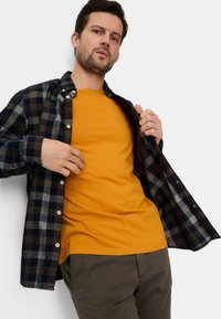 Modell trägt ein marineblaues und braunes kariertes Hemd über einem leuchtend orangenen T-Shirt. Graue Hose mit lockerem Sitz und strukturiertem Stoff.