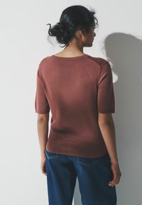 Krátký rukáv, vínový pletený svetr s žebrovaným lemem a kulatým výstřihem. Hladká textura a minimalistický design. Kombinován s modrými džínovými kalhotami.