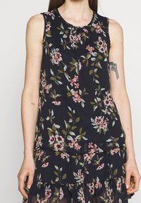 Top sans manches à motifs floraux avec un col rond en navy foncé, présentant des fleurs roses, blanches et vertes. Tissu léger avec des détails froncés.