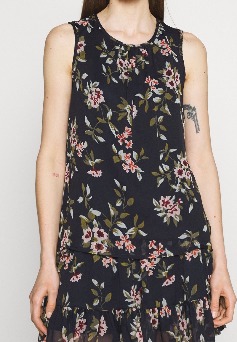 Top sans manches à motifs floraux avec un col rond en navy foncé, présentant des fleurs roses, blanches et vertes. Tissu léger avec des détails froncés.