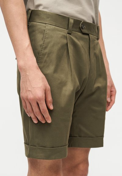 Άνδρας που φοράει καλοραμμένα κοντά παντελόνια σε ελιά πράσινο με πιέτες μπροστά και γυρισμένα τελειώματα, σε ουδέτερο φόντο.