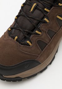 Brun vandresko med en struktureret nubuck-overdel, mesh-detaljer og kontrasterende gule accenter. Modellen har en robust gummisål for godt greb.