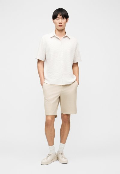 Νέος άνδρας στέκεται με τα χέρια στις τσέπες, φορώντας ένα λευκό υφασμάτινο πουκάμισο με κοντά μανίκια, μπεζ σορτς, λευκές κάλτσες και μπεζ αθλητικά παπούτσια.