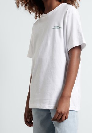 Person med krøllet hår iført en hvid Element Skateboard Co. t-shirt og lyseblå jeans, stående mod en ensfarvet baggrund.