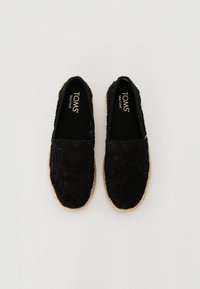 Et par svarte TOMS slip-on-sko med teksturert tekstiloverdel og beige jute-såler, sett ovenfra på en hvit bakgrunn.