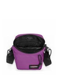 Sac bandoulière en nylon violet avec une fermeture éclair noire et une doublure intérieure. Comprend un patch logo et une sangle d'épaule réglable.