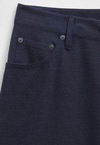 Donkerblauw denimstof met een voorzak met een knoopdetail, voorzien van riemlussen en stiksels langs de randen.