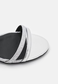 Sandale noire et blanche avec deux brides argentées brillantes et une semelle lisse et brillante blanche. La semelle est plate et texturée.