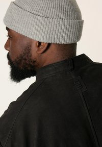 Schwarze Lederjacke mit hohem Kragen, ausgestattet mit einer strukturierten Oberfläche und einem gesteppten Logodetail. Eine hellgraue Strickmütze wird oben getragen.
