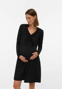 Robe de maternité noire à manches longues avec un col en V, des détails froncés au niveau du buste et une jupe fluide. Confectionnée dans un tissu texturé.