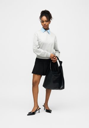 Femme portant un sweat-shirt gris par-dessus une chemise à col bleu, une jupe plissée noire, des talons noirs, et tenant un grand sac à main noir texturé.