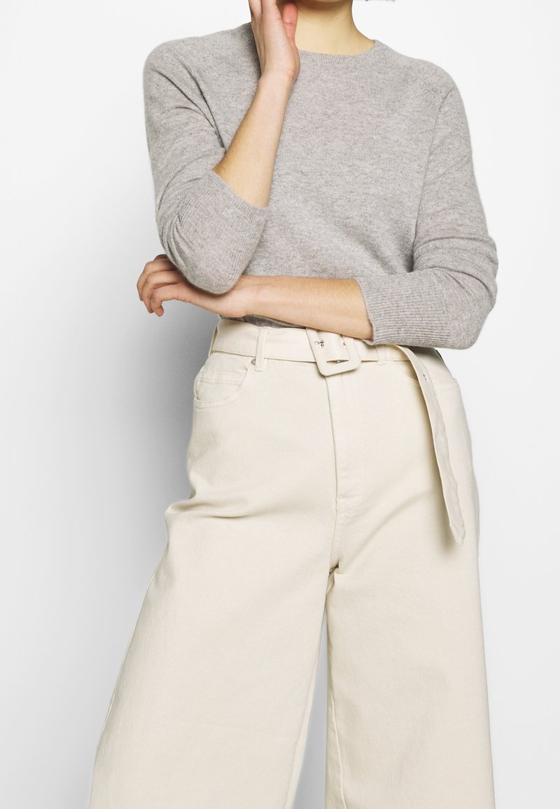 Femme portant un pull gris clair à manches longues, rentré dans un pantalon crème taille haute à jambes larges avec une ceinture en tissu assortie.