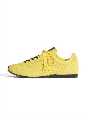 Żółty niski sneaker z czarną podeszwą i czarnymi przelotkami, wykonany z gładkiej i teksturowanej tkaniny, zawiązany żółtymi sznurowadłami, pokazany z boku.