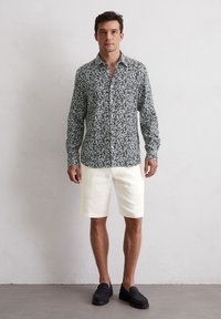 Herren Langarmhemd in Dunkelgrün mit floralem Muster, kombiniert mit weißen Shorts und schwarzen Slippern, vor einem schlichten Hintergrund stehend.