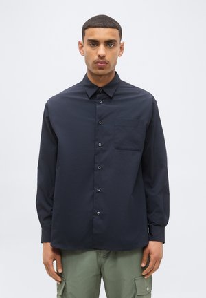 Uomo con capelli corti che indossa una camicia blu navy scura con bottoni e pantaloni cargo chiari, in piedi davanti a uno sfondo chiaro e uniforme.