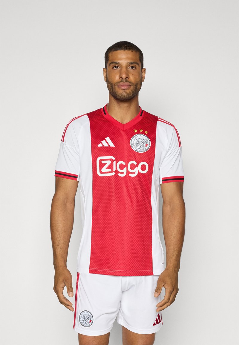 Maillot de football Ajax avec des couleurs rouge et blanche, un design col en V, une texture en mesh et un logo avec trois étoiles au-dessus.