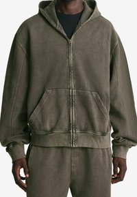 Sudadera con capucha y cremallera color verde oliva, confeccionada con un tejido suave, que cuenta con una capucha grande, bolsillos delanteros y puños y dobladillo acanalados.