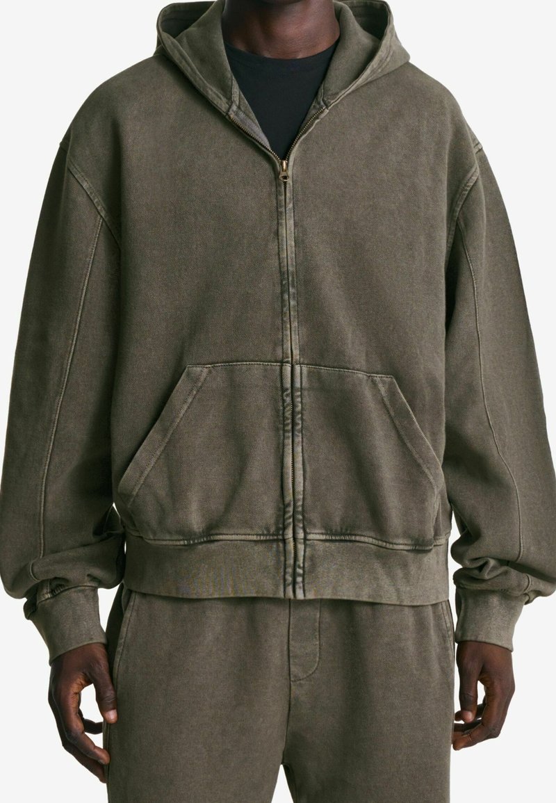 Sudadera con capucha y cremallera color verde oliva, confeccionada con un tejido suave, que cuenta con una capucha grande, bolsillos delanteros y puños y dobladillo acanalados.