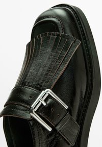 Zwarte leren schoen met een franjesdecoratie op de neus en een zilveren gesp op de band. Soepele textuur en afgeronde vorm.