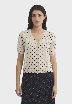 Femme portant un haut beige à manches courtes boutonné avec des pois noirs et une jupe noire à petits points blancs, debout devant un fond uni.