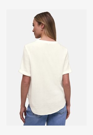 Vrouw met lang haar, gekleed in een gestructureerd wit kortermig topje en een blauwe spijkerbroek, gezien van achteren tegen een effen achtergrond.