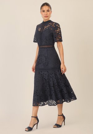 Navy lace midi dress with a high collar, short sleeves, floral pattern, and fitted waist. Paired with open-toe navy heels.