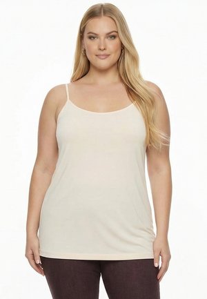 Kvinde med langt blond hår iført en beige ærmeløs top og mørke bukser, stående mod en ensfarvet hvid baggrund.