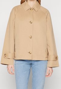 Beige bavlněná bunda s límcem, šesti velkými knoflíky a širokými rukávy. Má boční kapsy a rovný lem. Nosit na modrých džínách.