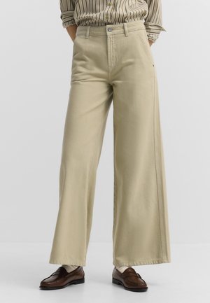 Person, die beige Hosen mit weitem Bein, braune Leder-Loafer, weiße Socken und ein in die Hose gestecktes gestreiftes Hemd mit hochgekrempelten Ärmeln trägt.