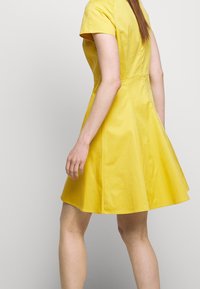 Vrouw draagt een felgeel jurkje met korte mouwen en een knie-lengte, staand tegen een effen lichtgrijze achtergrond.