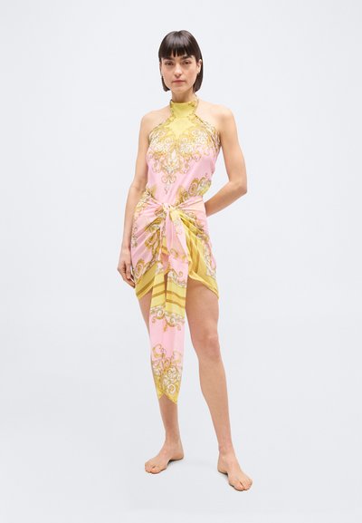 Γυναίκα που στέκεται ξυπόλητη, φορώντας ροζ και κίτρινο φόρεμα με σχέδιο, με δέσιμο υφάσματος στη μέση, πάνω σε απλό λευκό φόντο.