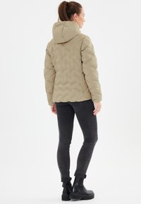 Beige, kviltad pufferjacka med huva, med ett texturerat mönster, bärs tillsammans med mörkgrå skinny jeans och svarta ankelkängor.