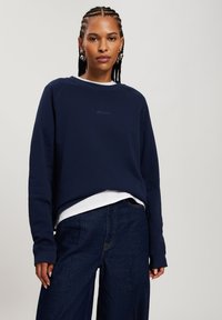 Granatowa bluza z okrągłym dekoltem i ściągaczami, noszona na białej koszuli. Posiada subtelne logo na piersi.