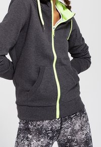 Sudadera gris con cremallera y forro interior de color verde neón, cuenta con bolsillos delanteros, llevada sobre leggings de patrón negro y gris.