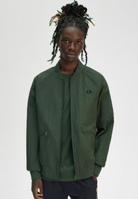 Bomber in verde scuro con una texture liscia, tasche con zip e polsini a coste. Indossato su una camicia verde coordinata.