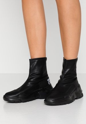 Czarne skórzane botki za kostkę z grubymi podeszwami, noszone na gołe nogi, z elastycznymi panelami po bokach i białym logo na obszarze pięty.