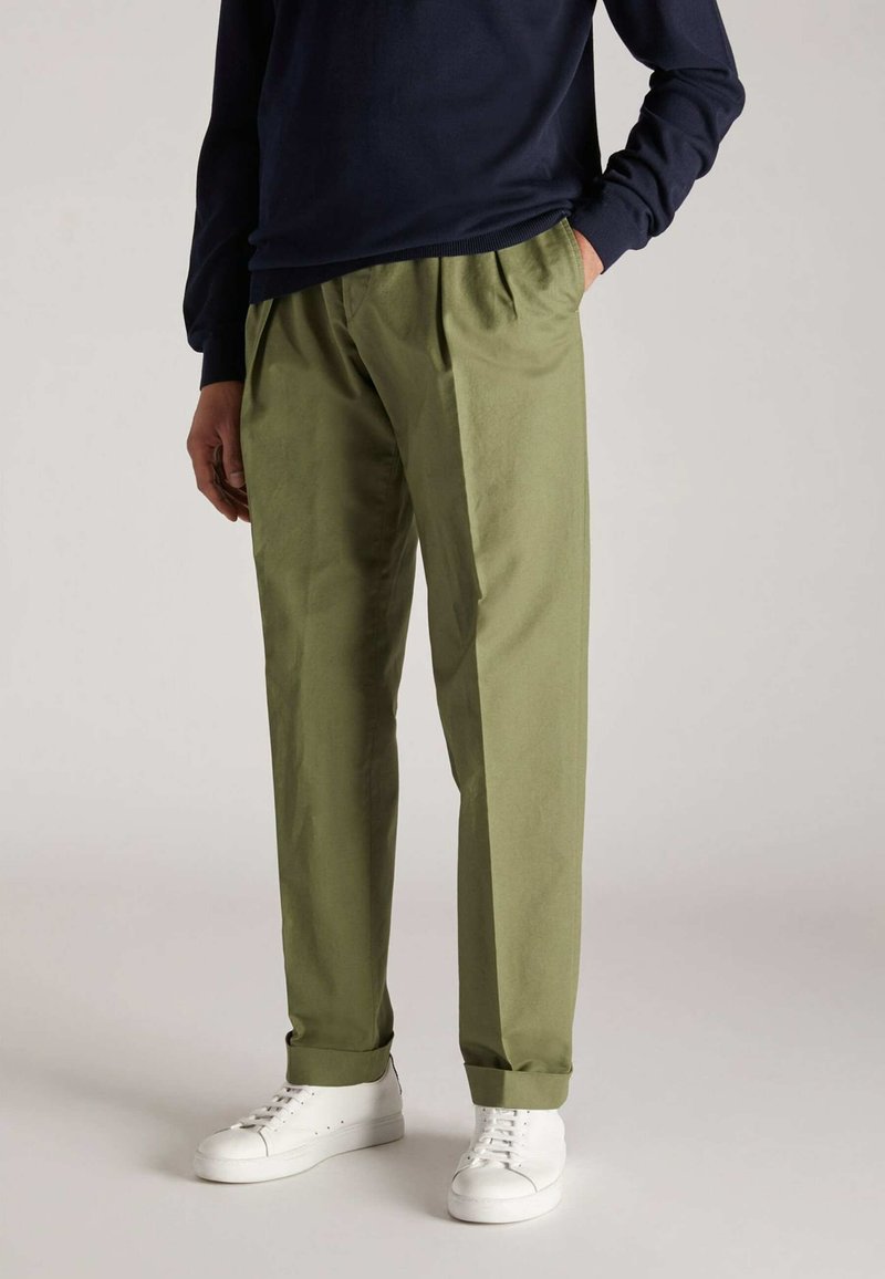 Olivgrüne, plissierte Hose mit lockerer Passform, aus glattem Stoff gefertigt, kombiniert mit weißen Sneakers und einem marineblauen Langarmshirt.