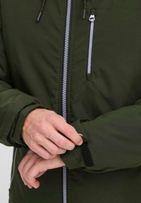 Giacca verde scuro con zip frontale, zip argentata e polsini regolabili con velcro. Presenta una texture di tessuto liscia e elegante.